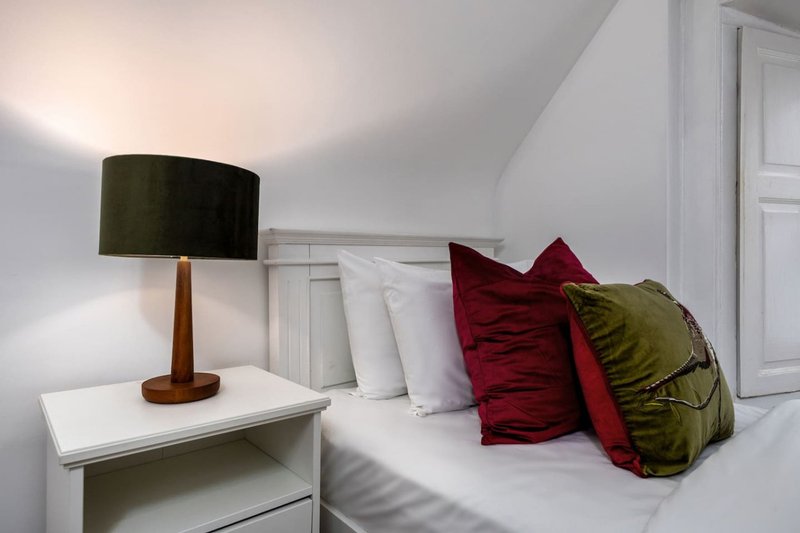 Bedroom with white bedside table, wooden lamp, red and green cushions on bed