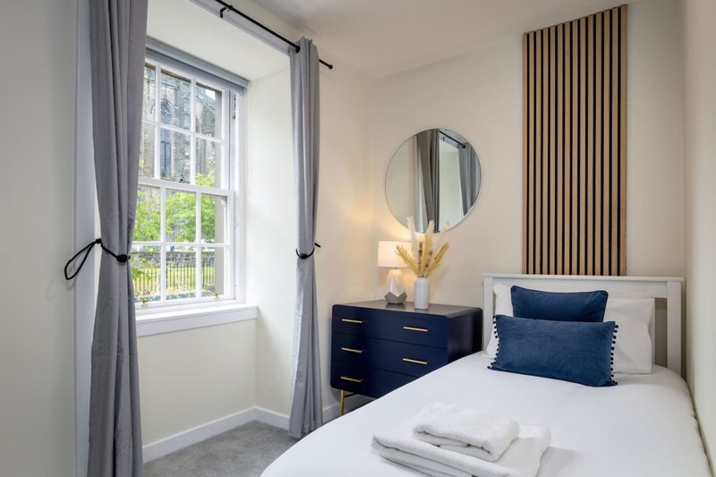 Modern bedroom with double bed, blue pillows, dark dresser, round mirror and window with grey curtains