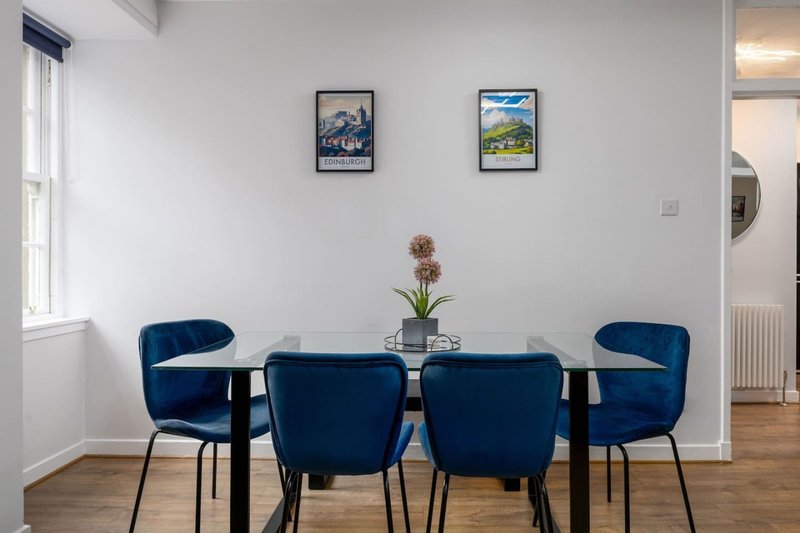 Dining room with glass table, four blue upholstered chairs, and framed artwork on walls