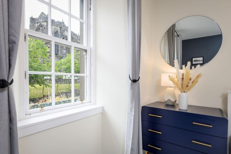 Bright bedroom corner with large window, dark blue dresser, round mirror and dried flowers in vase