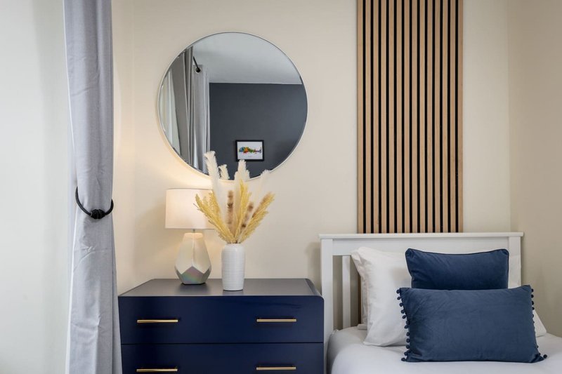 Modern bedroom with dark dresser, round mirror, white bed with blue pillows and gold décor accents