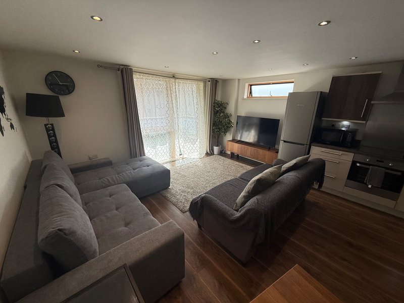 Open-plan living room with two grey sofas, TV, kitchen area, and glass doors to garden