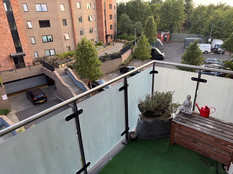 Rooftop terrace with frosted glass railing, potted plants, red watering can, and urban courtyard view