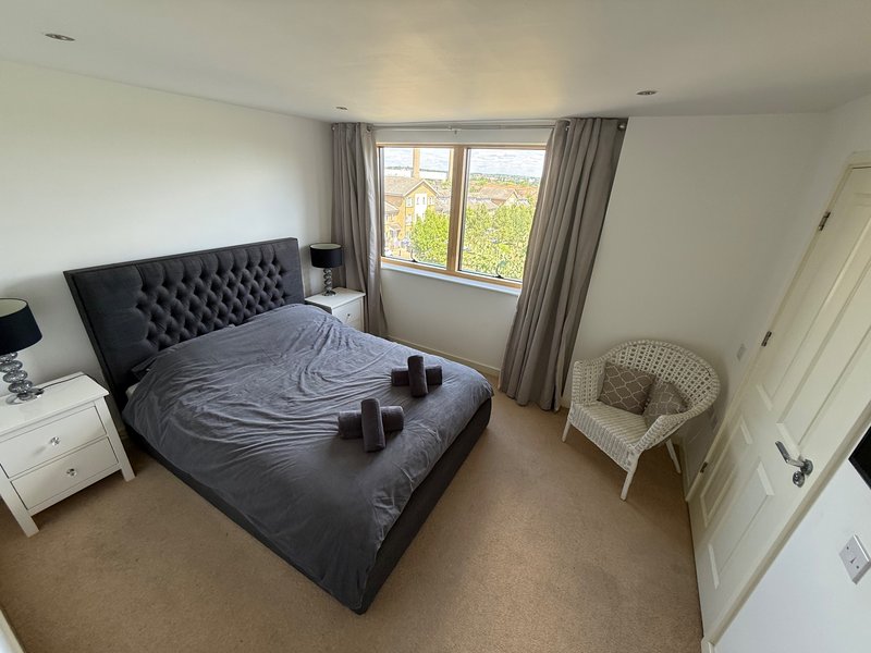Spacious bedroom with upholstered bed, wicker chair, large windows with grey curtains, white furniture