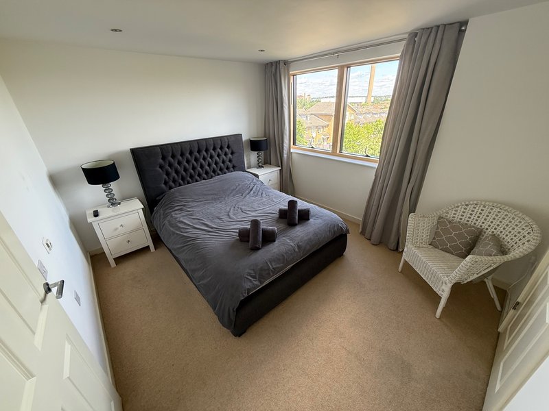 Bright bedroom with black upholstered bed, white nightstand, chair, and large window with grey curtains