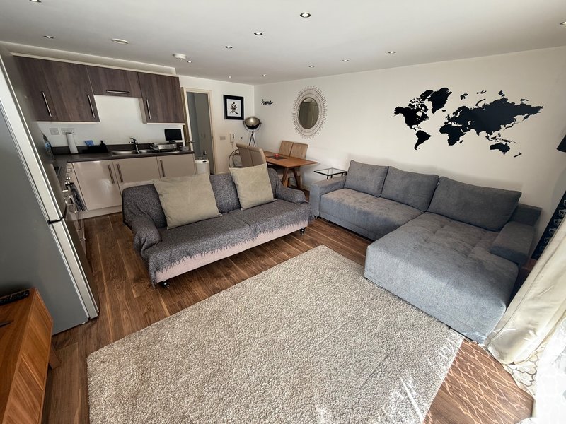 Open-plan living room with grey corner sofa, wooden flooring, kitchen area, and world map wall art