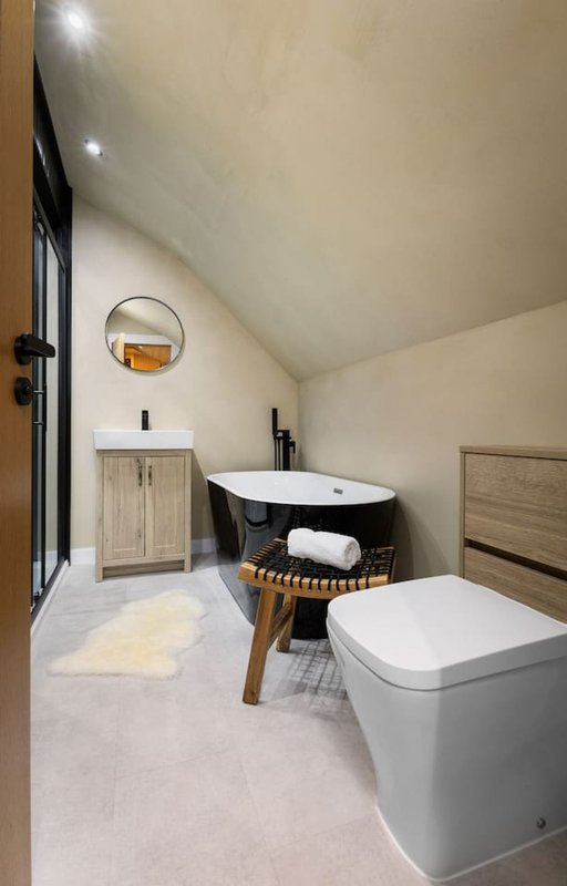 Modern bathroom with black freestanding bathtub, white toilet, wooden vanity, round mirror and skylight