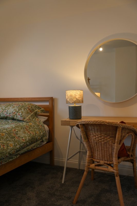 Bedroom with wooden bed, patterned bedding, round mirror, desk with lamp and wicker chair