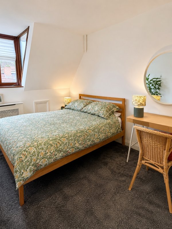 Bright bedroom with wooden bed, patterned bedding, desk, round mirror and window.