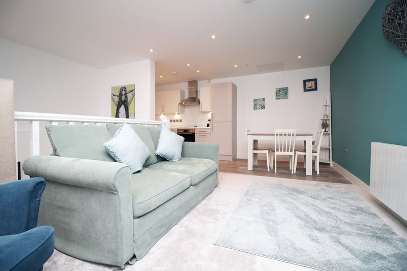 Open-plan living room with gray sofa, dining table, kitchen, and teal accent wall
