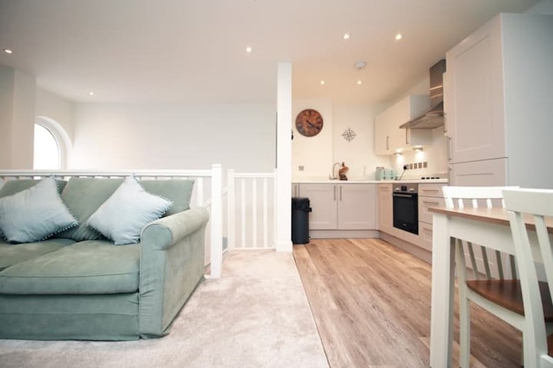 Open-plan living space with green sofa, white kitchen, wooden flooring and decorative clock