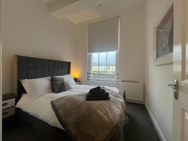 Modern bedroom with black headboard, double bed, bedside lamps, window with roller blind and city view