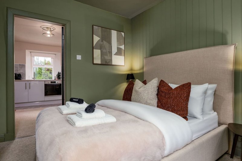 Modern bedroom with double bed, burgundy pillows, green walls and open-plan kitchen view