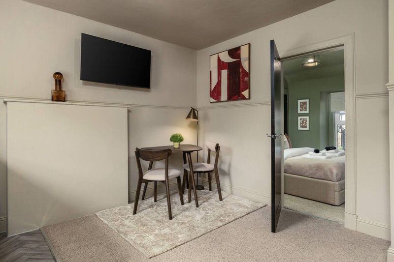 Modern studio apartment with wall-mounted TV, small dining table, and open bedroom visible through doorway