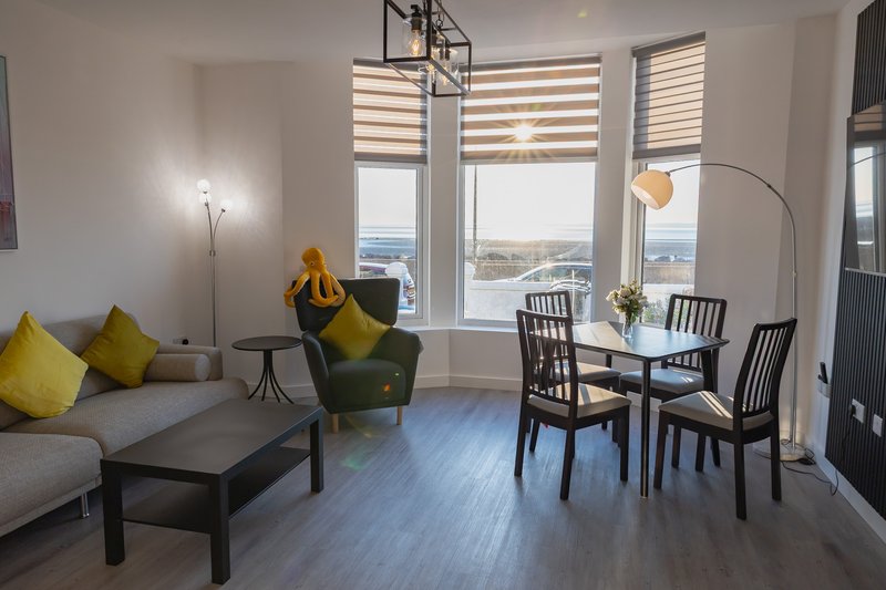 Spacious living area with gray sofa, yellow cushions, green armchair, dining table and large windows overlooking city.