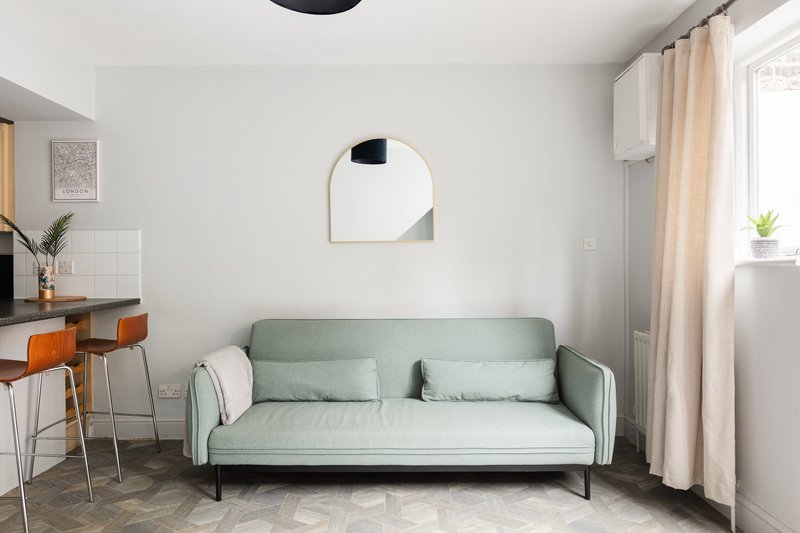 Modern living room with sage green sofa, round mirror, desk with orange chairs, and window with cream curtains