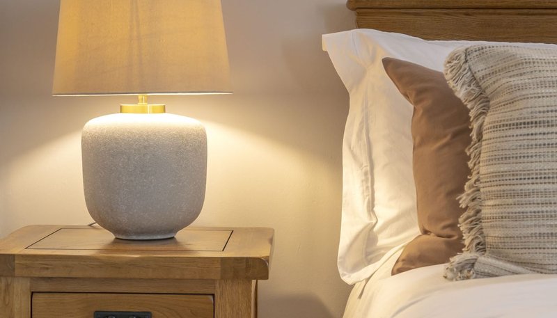 Bedroom bedside table with ceramic lamp and wooden nightstand, adjacent to bed with pillows