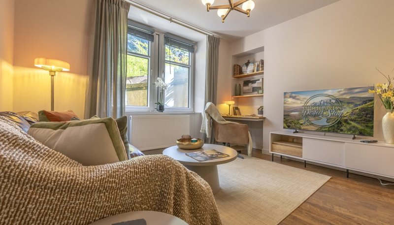 Bright living room with sofa, TV unit, desk area, and large windows overlooking garden