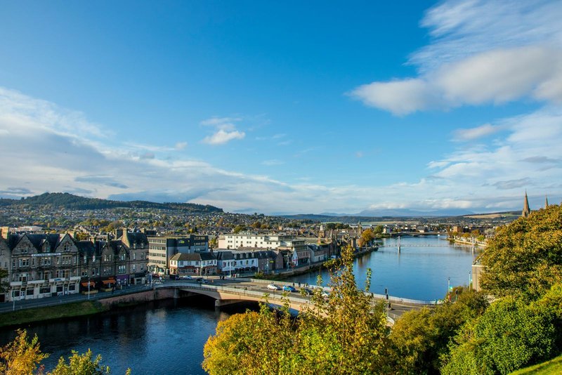 Riverside town view with bridge, buildings and forested hills under blue sky