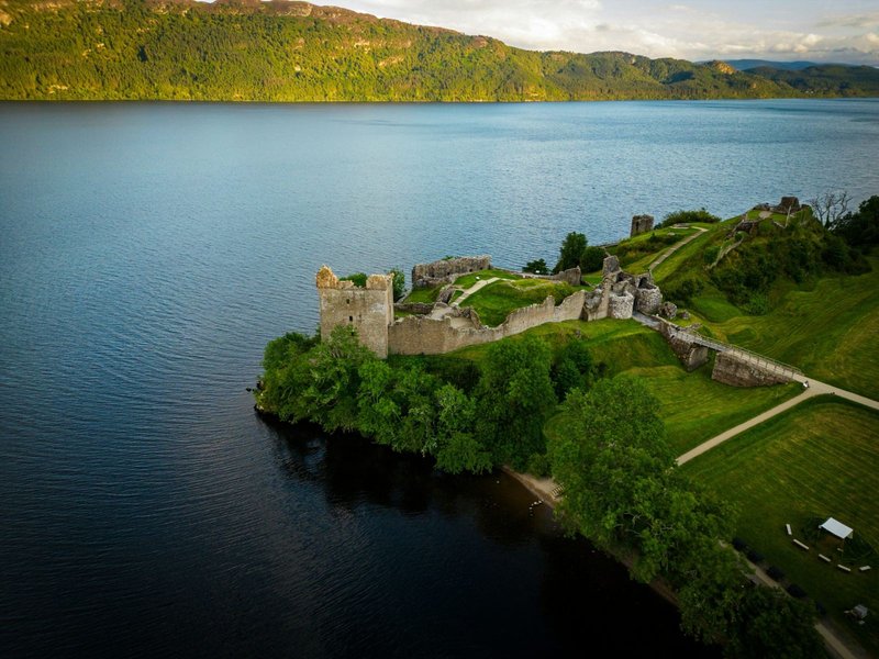 Aerial view of lakeside property with ruined stone castle on grassy peninsula surrounded by water