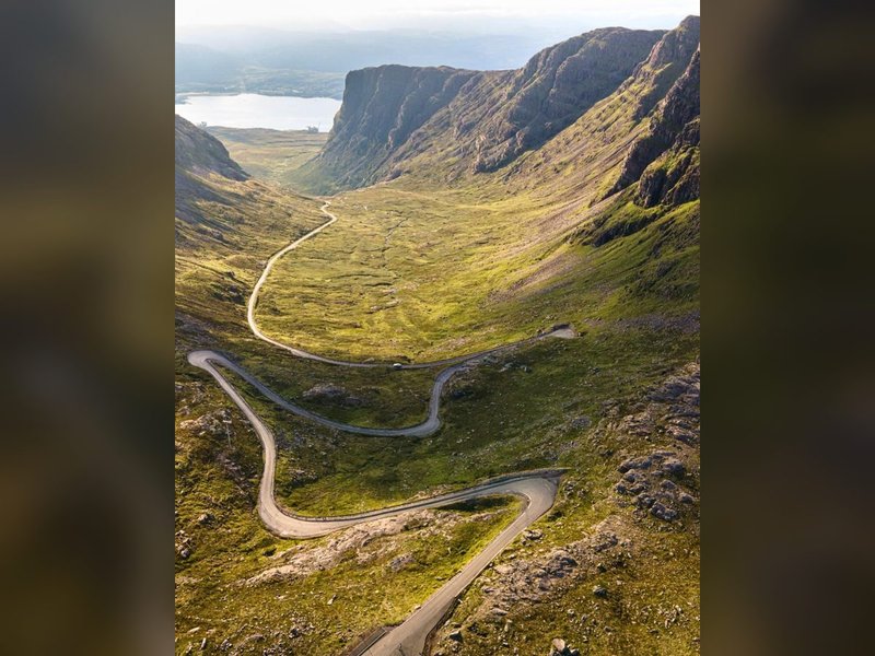 Aerial view of winding mountain road through green valley with distant lake and steep cliffs