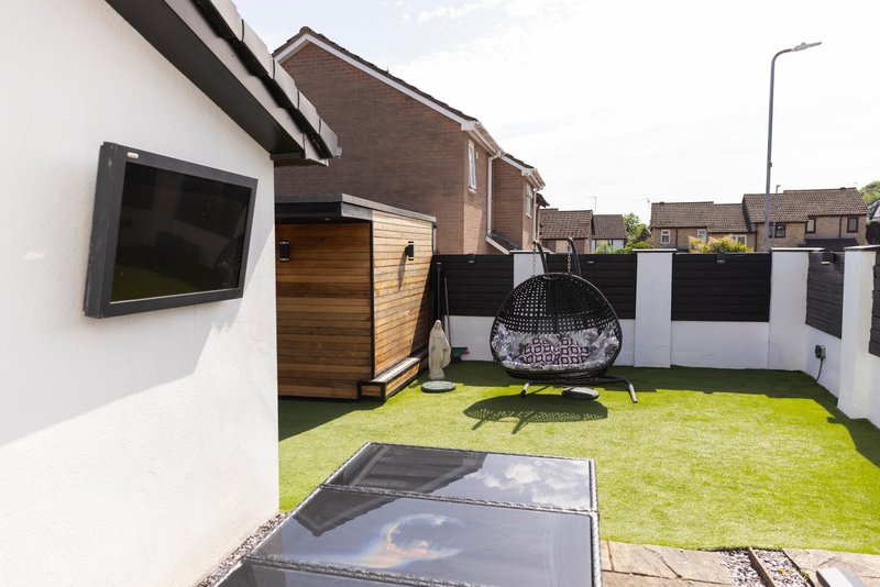 Modern house exterior with paved patio, artificial grass garden, black parasol, and wooden outbuilding