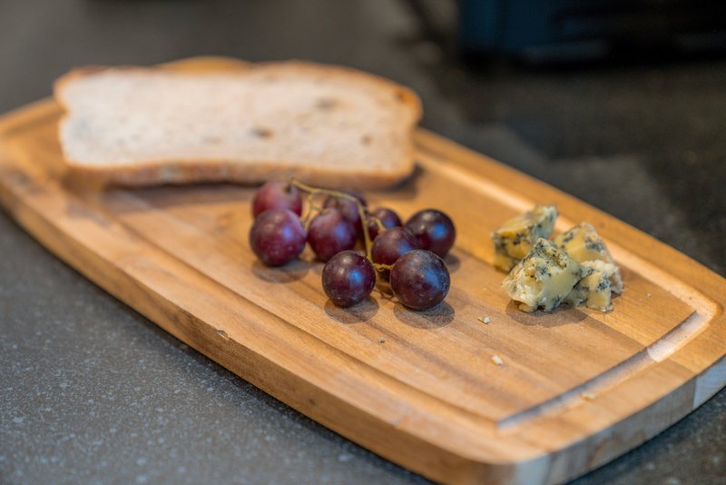 Wooden serving board with bread, grapes, and cheese on dark surface