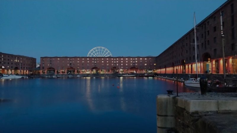 Waterfront view of historic dock buildings at dusk with illuminated Ferris wheel in background