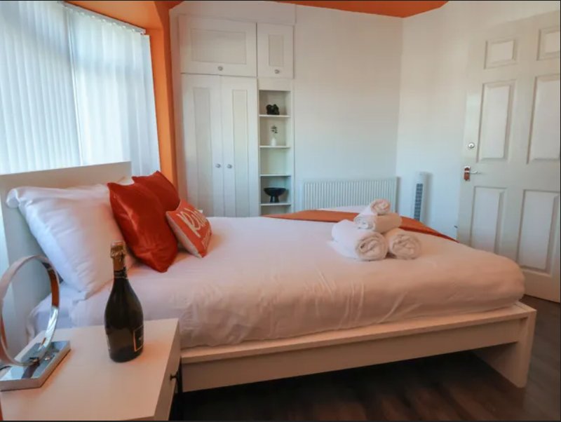 Bright bedroom with double bed, red cushions, wine bottle, and white fitted wardrobes