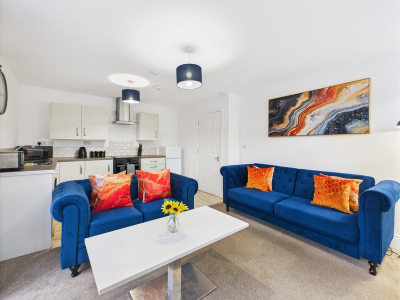 Open-plan living room with blue sofas, white coffee table, kitchen, and wall art