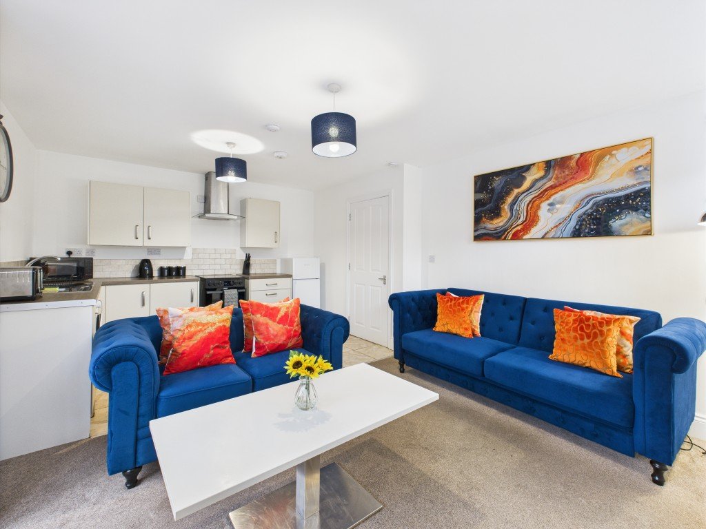Open-plan living area with blue sofas, white coffee table, kitchen, and abstract wall art