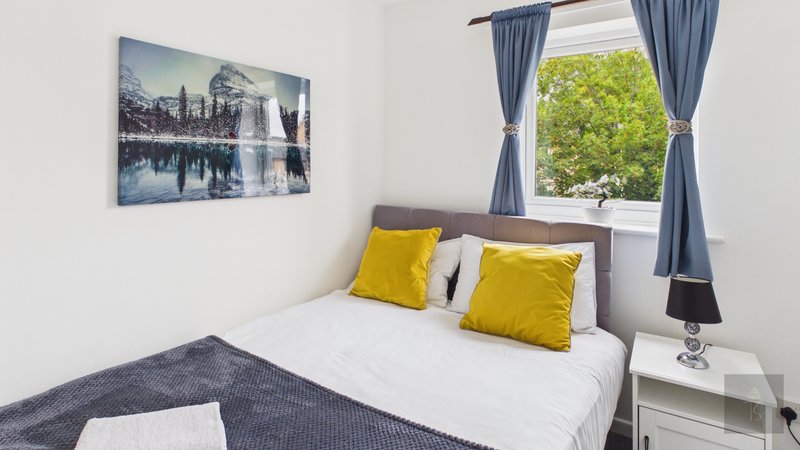 Bright bedroom with double bed, yellow cushions, framed landscape artwork, and window with garden view