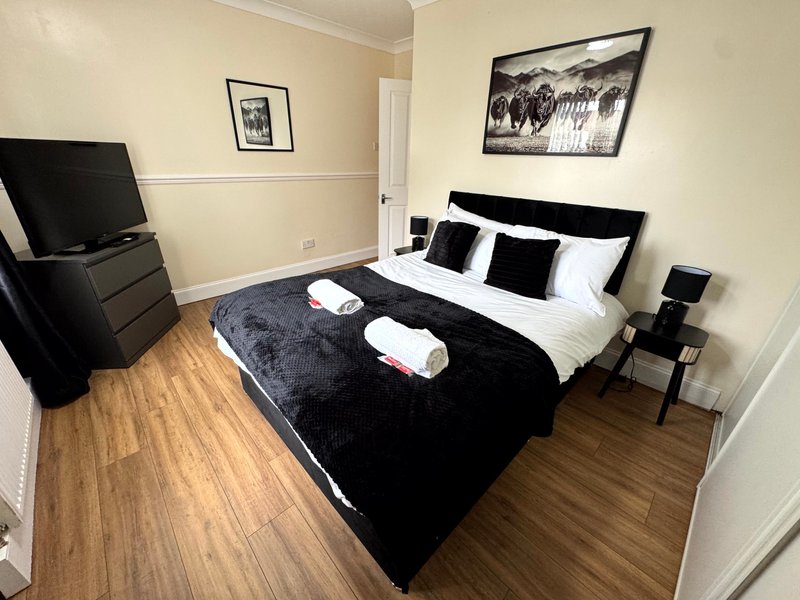 Modern bedroom with double bed, black headboard, white pillows, folded towels, wooden flooring and TV