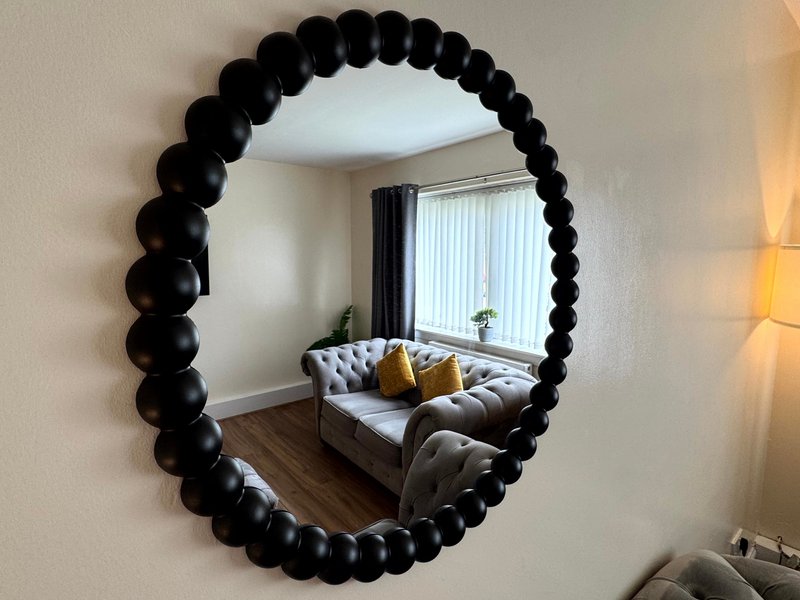 Modern living room with grey sofa, yellow cushions, window with curtains, reflected in large black beaded mirror