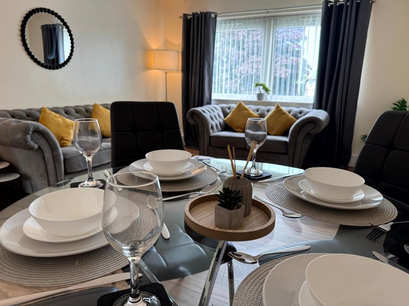 Dining table set for four with place settings, glasses, and centerpiece; living room with sofas visible in background