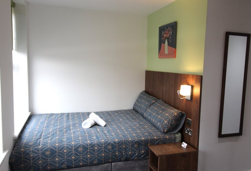 Compact bedroom with double bed, geometric patterned bedding, wooden headboard with integrated lighting, and green accent wall