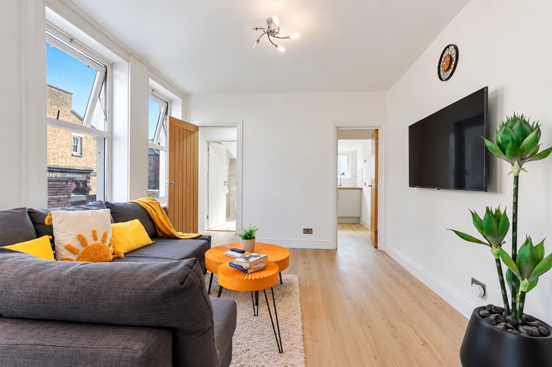 Bright living room with grey sofa, orange cushions, wall-mounted TV, and potted plants