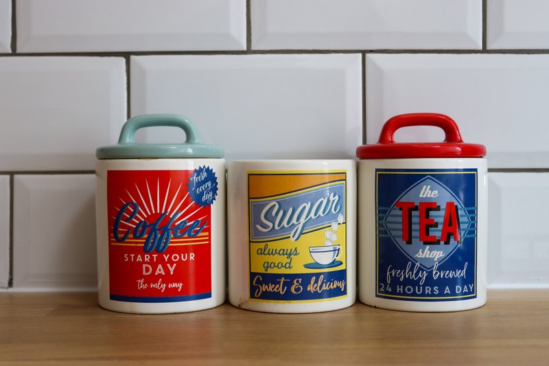 Three branded kitchen storage containers with colorful retro-style labels on white subway tile backsplash