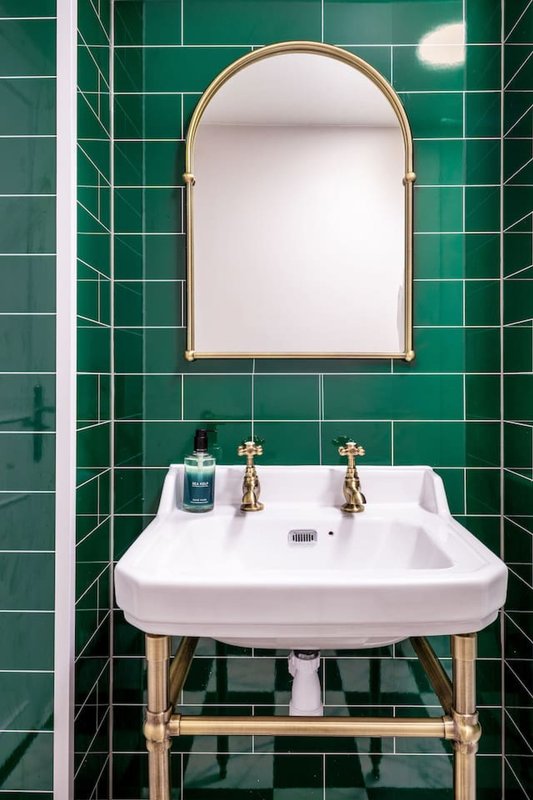 Modern bathroom with emerald green tile walls, pedestal sink with brass fittings and arched mirror
