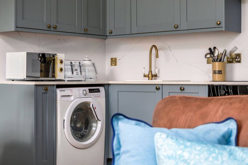 Modern kitchen with gray cabinetry, white washing machine, brass tap and blue cushions visible