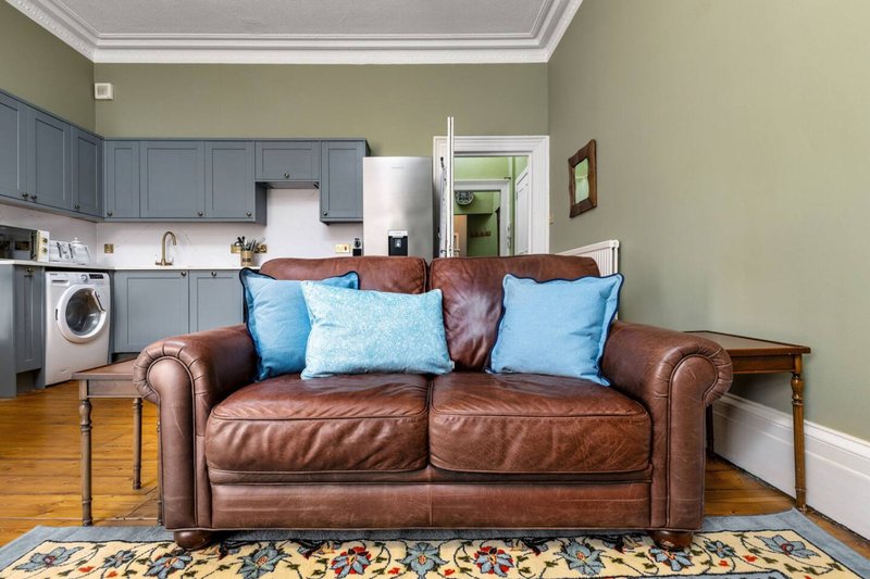 Open-plan living area with brown leather sofa, blue cushions, blue cabinetry kitchen, and wooden flooring