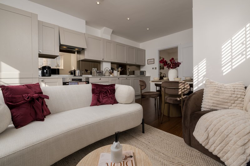 Open-plan living area with cream sofa, burgundy cushions, kitchen, and natural light from windows