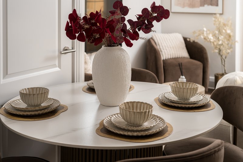 Modern dining table set with place settings, white vase with burgundy flowers, and upholstered chairs