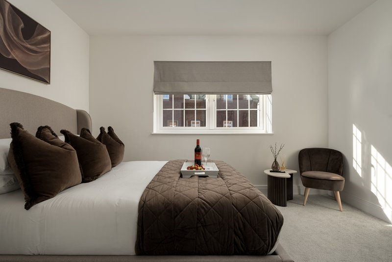 Modern bedroom with white bed, brown cushions, armchair, and large window with grey blind
