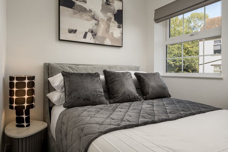 Modern bedroom with grey quilted bedding, dark cushions, framed abstract art, and large windows