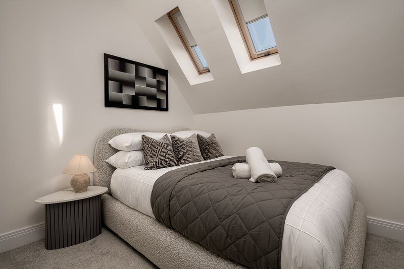 Modern bedroom with grey upholstered bed, white pillows, dark quilt, skylight windows and wall art