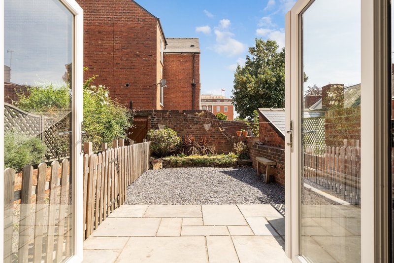 Outdoor patio area with gravel and paving, wooden fencing, brick buildings and trees visible