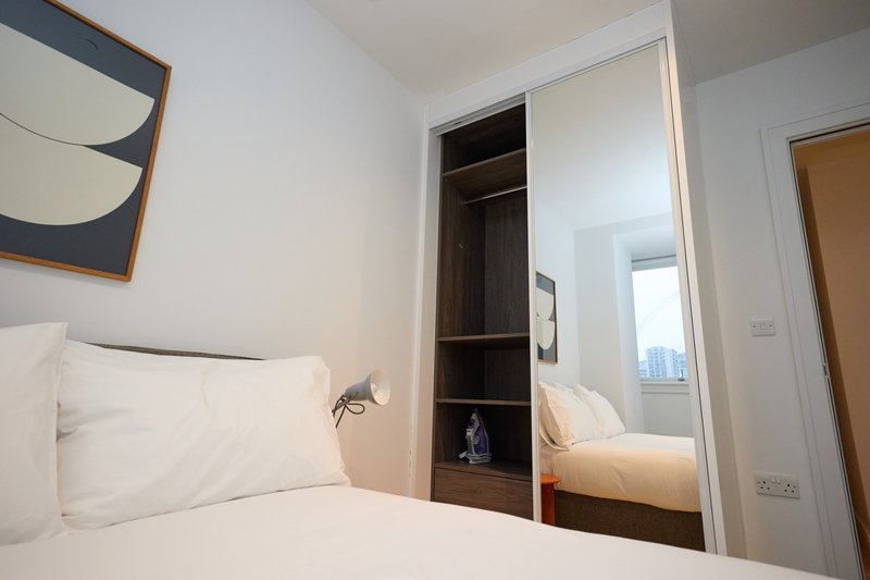 Compact bedroom with white bedding, built-in wardrobe, mirror, and window with city view
