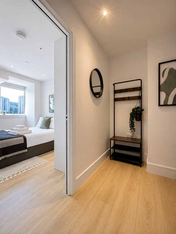 Modern bedroom entryway with wooden flooring, wall-mounted mirror, coat rack, and view into bedroom beyond