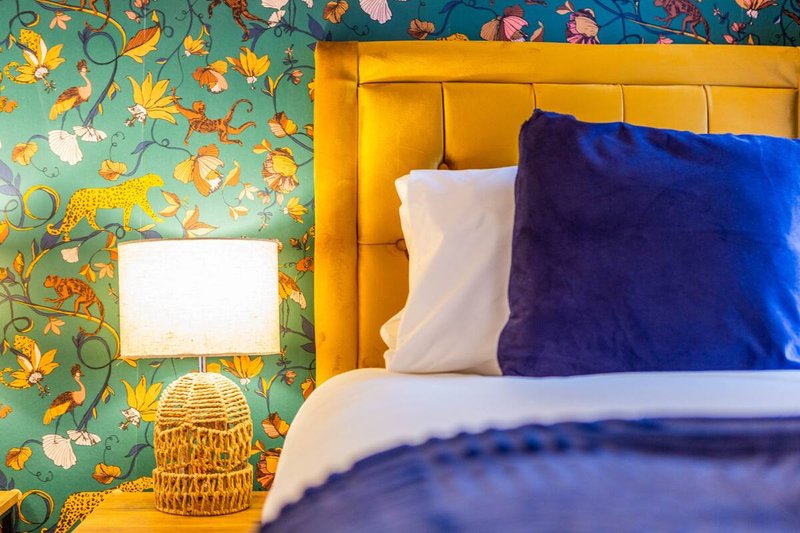 Bedroom with yellow upholstered headboard, colorful floral wallpaper, blue and white bedding, wicker lamp